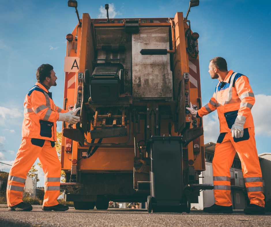 Dva popeláři v oranžovém pracovním oděvu nakládající popelnice do zádi odpadového vozu